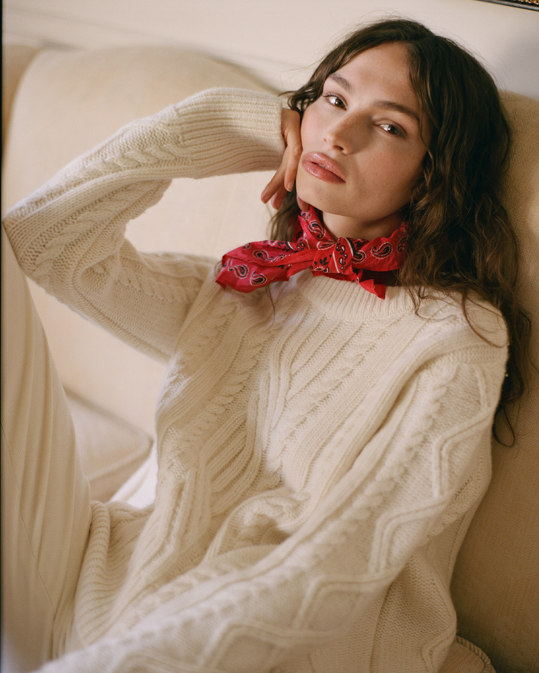 brunette wearing a cable knit cream jumper with a red patterned neck scarf, on a cream sofa.